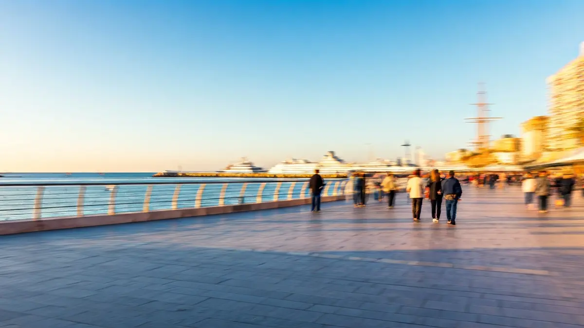 Imagen de un paseo peatonal moderno junto al puerto de Almería, con personas caminando y barcos al fondo.