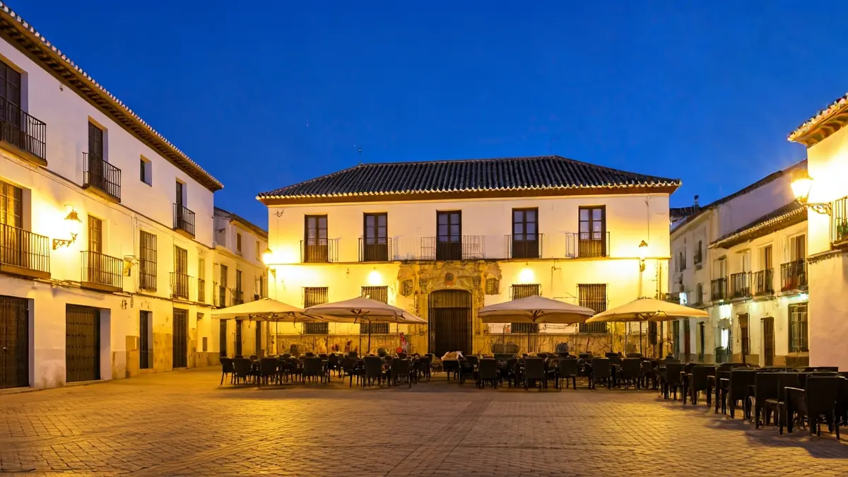 Imagen de una plaza en un pueblo andaluz al atardecer, con terrazas llenas de gente y edificios históricos iluminados.