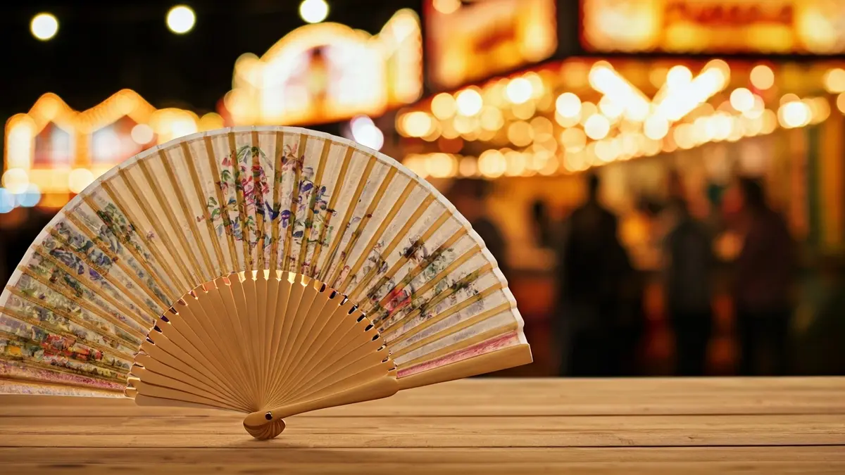 Imagen de un abanico tradicional en una mesa de madera, con luces de feria borrosas al fondo.