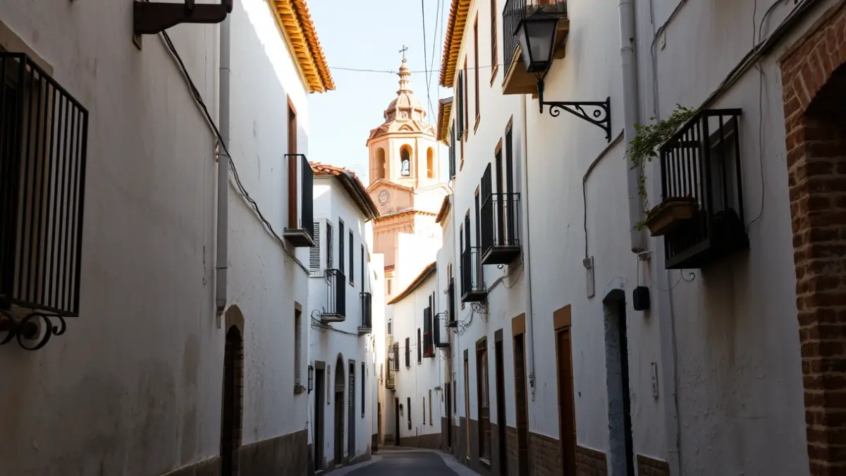 Imagen de una calle tradicional andaluza con edificios encalados y una torre de iglesia al fondo.