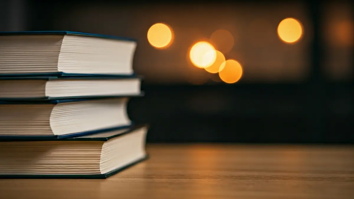 Imagen genérica de libros apilados en una mesa de madera, con luz cálida.