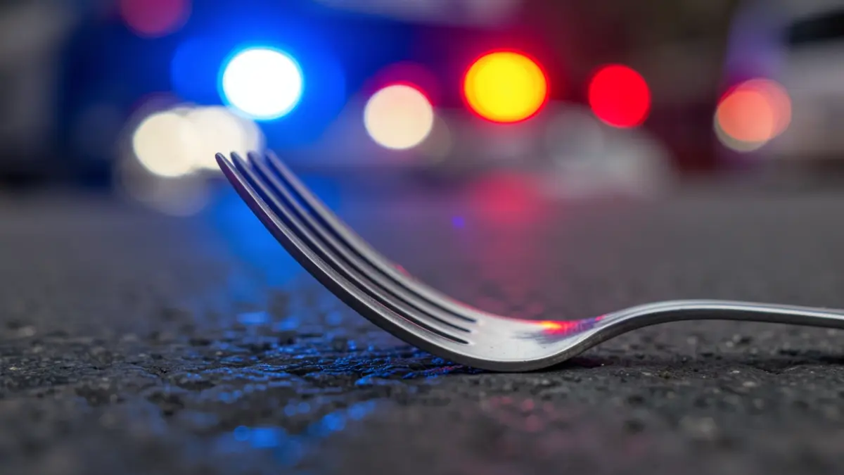 Close-up of a metal fork with a slight bend on a dark surface, with blurred blue and red emergency lights in the background.