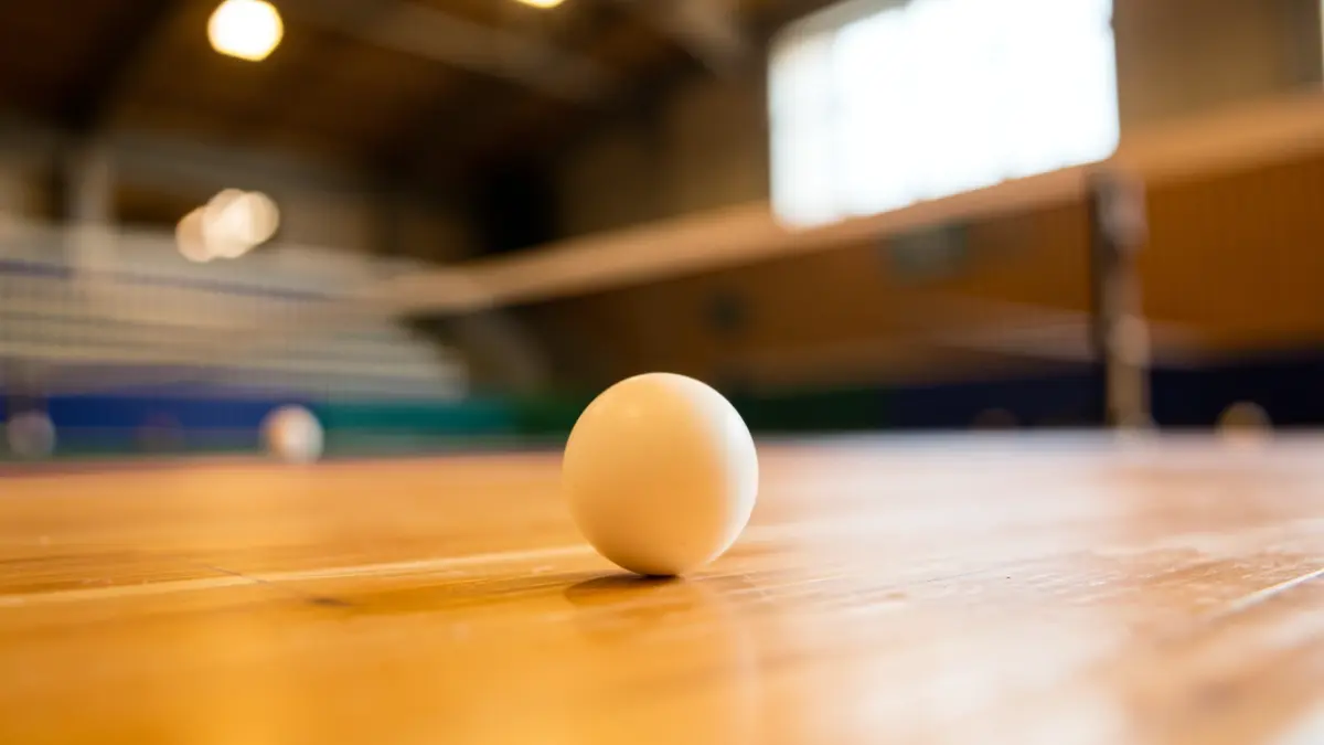 Close-up of a table tennis ball on a wooden table, with a blurred background of a sports hall and net.