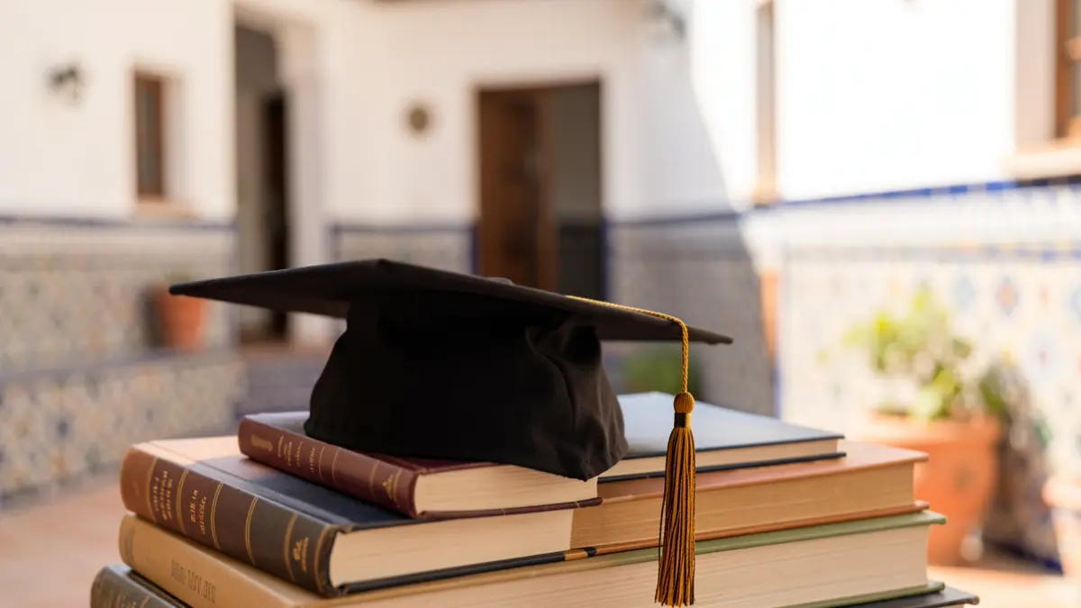 Imagen genérica de un birrete de graduación sobre libros, con un patio andaluz al fondo.