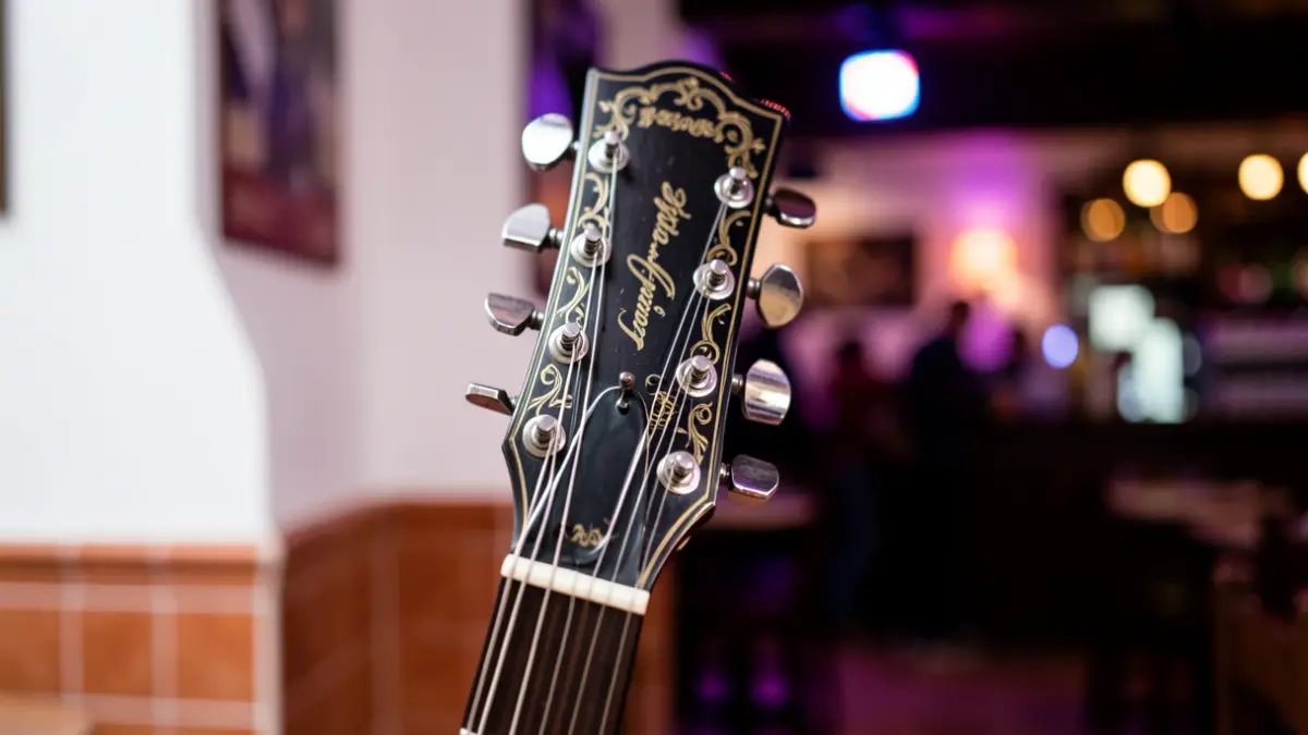 Close-up of a guitar headstock with intricate detailing, set against a blurred background of a dimly lit pub interior with subtle stage lighting, evoking a rock and roll atmosphere.