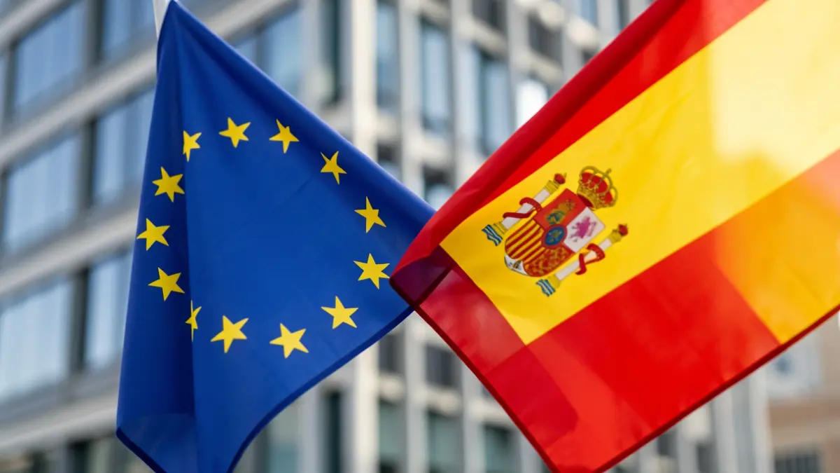 Spanish and European Union flags intertwined in front of a modern office building.
