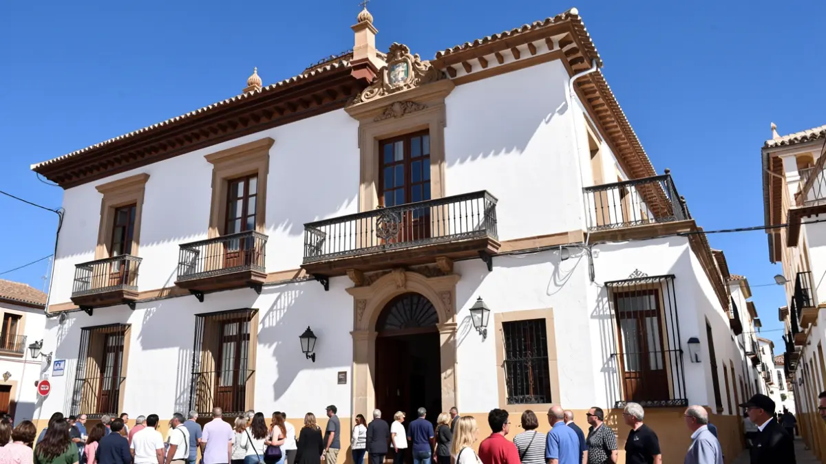 Fachada de un ayuntamiento andaluz con balcón ornamentado y paredes blancas, bajo el sol brillante del mediodía.