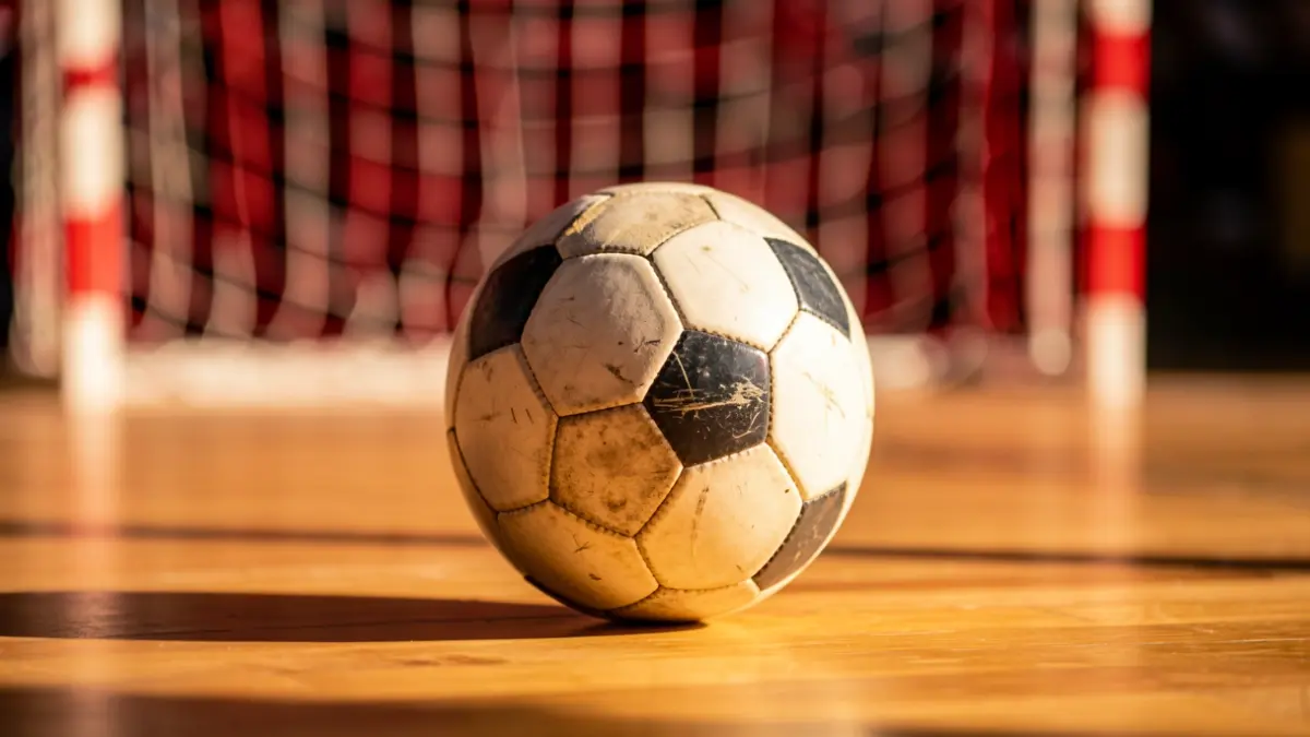 Primer plano de un balón de fútbol sala sobre una cancha de madera con redes de portería borrosas al fondo.