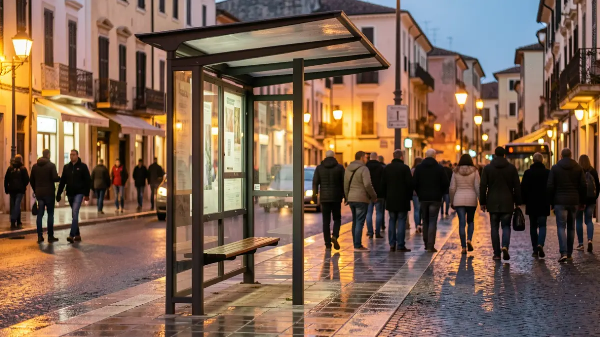 Bus stop shelter in a Mediterranean town at dusk, with warm streetlights illuminating the wet pavement and blurred building facades in the background.